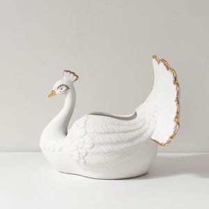 May include: A white ceramic peacock-shaped planter with gold detailing. The peacock has a long neck, detailed head, and a fanned tail. The planter is designed to hold plants or flowers, adding a decorative touch to any room.