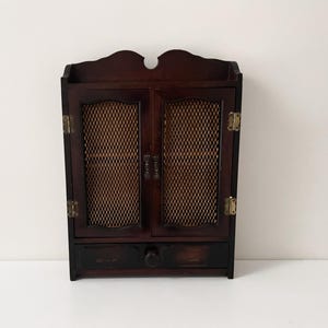 Peut inclure: Une armoire murale en bois brun foncé dotée de deux portes avec des panneaux en maille métallique dorée, des charnières couleur laiton et un tiroir inférieur unique avec un bouton rond en bois. Le haut présente un bord incurvé.