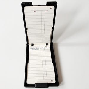 Puede incluir: Libreta de direcciones de cuero negro, abierta para revelar p&aacute;ginas rayadas. El libro tiene forma rectangular con una tapa con bisagras y un cierre. Las p&aacute;ginas son blancas con l&iacute;neas horizontales. Las iniciales "KL" son visibles.
