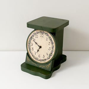 May include: A dark green wooden clock shaped like a kitchen scale. The round clock face features a cream background with black numbers from 1 to 12, black hands, and a decorative border of small purple flowers. Gold painted accents decorate the base.
