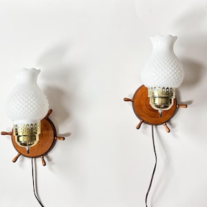 May include: A pair of vintage wall sconces. Each has a white, textured glass shade above a brass fitting, set on a wooden ship's wheel design. The sconces are connected to a black electrical cord. The background is a plain white wall.