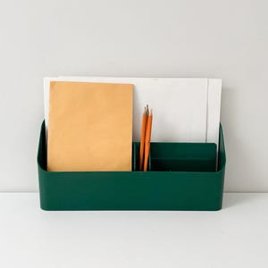 May include: A dark green desk tidy containing stationery. It holds a manila envelope, white paper, and orange pencils. The organiser sits on a white surface against a white wall.