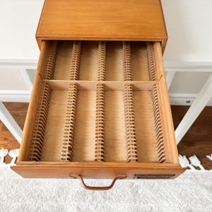 Puede incluir: Cajón de cubiertos de madera con múltiples ranuras para organizar utensilios. El cajón está abierto, revelando los divisores interiores. La madera tiene un tono cálido y natural, y el cajón está sobre un fondo neutro.