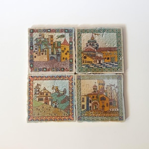 Op de afbeelding: Een set van vier vierkante keramische tegels met gedetailleerde illustraties van kastelen en stadsgezichten in middeleeuwse stijl. Elke tegel is omrand met een kleurrijk geometrisch patroon en toont getextureerde, veelkleurige architecturale scènes.