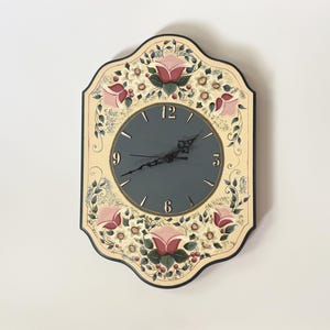 May include: A wall clock with a scalloped, cream-coloured frame featuring hand-painted pink roses, green leaves, and small white flowers. The central circular dial is dark grey with black hands and gold-coloured numerals at the 3, 6, 9, and 12 positions.