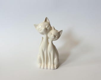 May include: A glossy off-white ceramic figurine depicting two stylized cats sitting side by side. The taller cat stands slightly behind the smaller one, with both figures featuring smooth, elongated necks, pointed ears, and closed eyes.