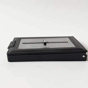 Puede incluir: Un calculador de fechas de cuero negro con un dial deslizante plateado. El calculador tiene un dise&ntilde;o rectangular y una peque&ntilde;a pesta&ntilde;a en el lateral. Est&aacute; sobre un fondo blanco. El calculador se utiliza probablemente para calcular fechas.