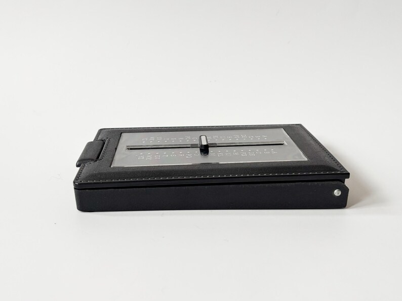 Puede incluir: Un calculador de fechas de cuero negro con un dial deslizante plateado. El calculador tiene un dise&ntilde;o rectangular y una peque&ntilde;a pesta&ntilde;a en el lateral. Est&aacute; sobre un fondo blanco. El calculador se utiliza probablemente para calcular fechas.