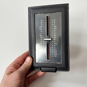 Puede incluir: Un directorio telef&oacute;nico vintage de cuero negro con un &iacute;ndice deslizante plateado. El &iacute;ndice tiene letras de la A a la YZ. El directorio est&aacute; sostenido por una mano sobre un fondo blanco.