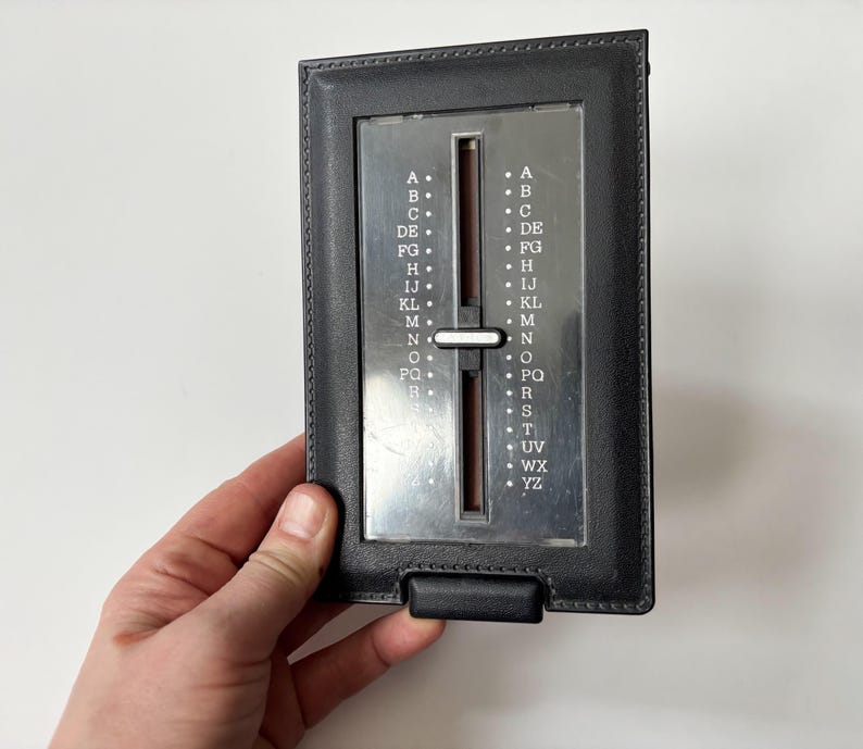 Puede incluir: Un directorio telef&oacute;nico vintage de cuero negro con un &iacute;ndice deslizante plateado. El &iacute;ndice tiene letras de la A a la YZ. El directorio est&aacute; sostenido por una mano sobre un fondo blanco.