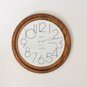 Peut inclure: Une horloge murale ronde avec un cadre en bois brun moyen et un cadran blanc. L'horloge présente des chiffres noirs aux lignes fines de un à douze, des aiguilles blanches, et le texte "Quarumatic" et "Seth Thomas" imprimé en noir.