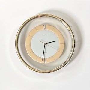 May include: A round wall clock with a gold-toned outer frame and a clear glass border. The center features a white dial with a beige ring, black Roman numerals at the twelve and six positions, black hands, and the text Verichron Quartz.