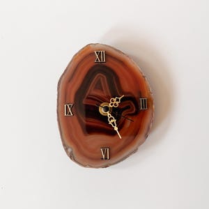 Puede incluir: Un reloj de pared hecho de una rodaja pulida de piedra ágata con bandas marrones y bronceadas. La esfera presenta manecillas de metal dorado y cuatro números romanos dorados aplicados en las posiciones de las doce, tres, seis y nueve.