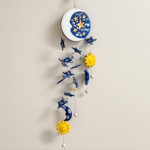 Op de afbeelding: Een witte keramische maan met een blauw en gouden sterrenontwerp hangt aan een touwtje met blauwe sterren, halve manen en gele zonnen.
