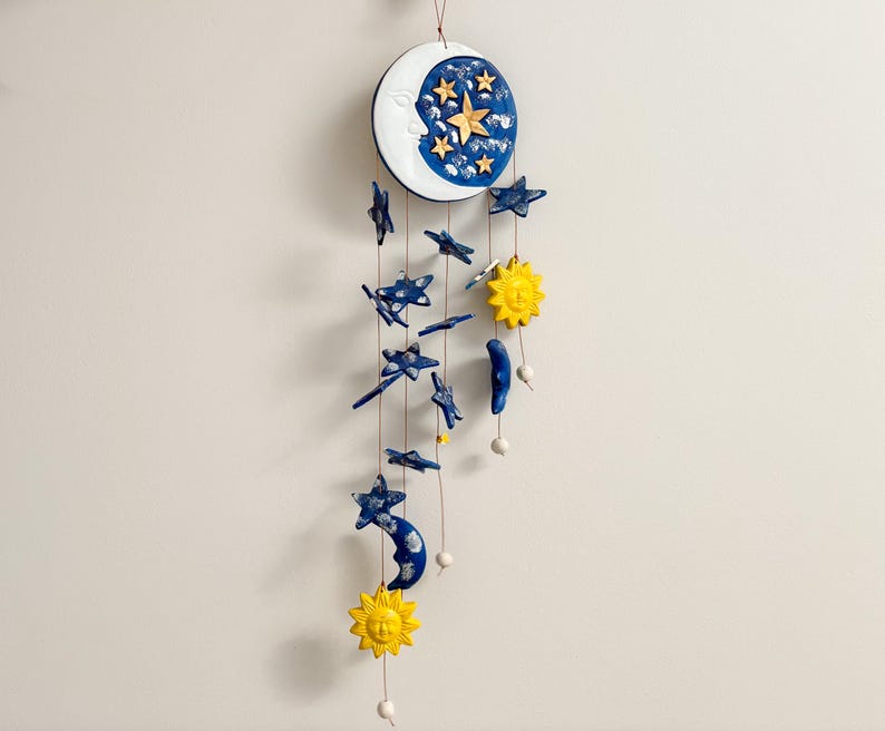 Op de afbeelding: Een witte keramische maan met een blauw en gouden sterrenontwerp hangt aan een touwtje met blauwe sterren, halve manen en gele zonnen.