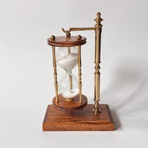 May include: An hourglass with a wooden base and top, brass supports, and a clear glass chamber filled with white sand. The hourglass is supported by a brass stand and is a decorative item.
