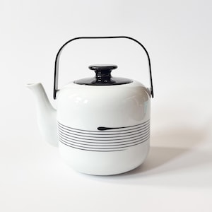 Może przedstawiać: Biały ceramiczny czajnik z czarnym uchwytem, pokrywką i poziomymi paskami. Czajnik ma zakrzywioną wylewkę i zaokrąglony korpus. Design jest minimalistyczny i nowoczesny, idealny do serwowania herbaty.