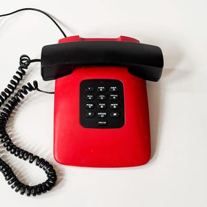 Peut inclure: Un téléphone rétro rouge avec un combiné et un clavier noirs. Le téléphone a un cordon noir enroulé et un cordon noir droit. Le clavier comporte des boutons noirs avec des chiffres et des lettres blancs.