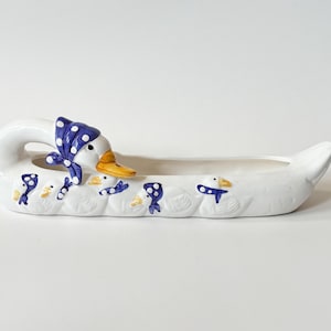 Può includere: Fioriera in ceramica bianca a forma di oca, con sciarpa blu a pois e becco giallo. La fioriera ha una forma ovale allungata con piccole figure di oca e dettagli blu. Un oggetto decorativo per la casa.