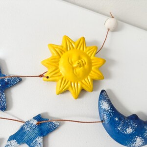 Op de afbeelding: Een gele zon met een gezicht, een blauwe halve maan en blauwe sterren aan een touwtje. De zon en de maan hebben een wit gespikkeld ontwerp.