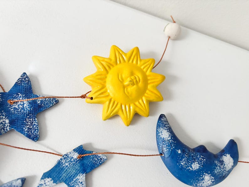Op de afbeelding: Een gele zon met een gezicht, een blauwe halve maan en blauwe sterren aan een touwtje. De zon en de maan hebben een wit gespikkeld ontwerp.