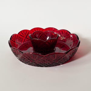 Puede incluir: Un juego de servir de cristal rojo intenso que consta de una bandeja grande y redonda con bordes festoneados y un patrón de corte de diamante, junto con un cuenco central más pequeño a juego. El cristal rubí translúcido tiene detalles geométricos.
