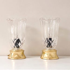 May include: Two clear glass table lamps with a ribbed design and a gold-colored base. The lamps have a diamond-shaped pattern at the bottom of the glass and a scalloped edge at the top. The lamps are decorative and provide ambient lighting.