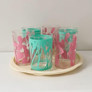 Pode incluir: Um conjunto de seis copos transparentes com desenhos rosa e turquesa, dispostos numa bandeja oval creme. Os copos apresentam formas rosas abstratas e detalhes turquesa.