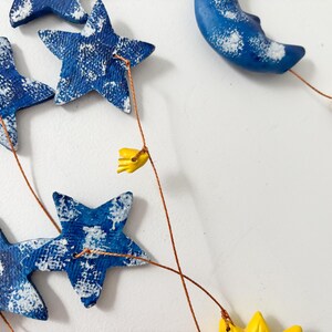Op de afbeelding: Een blauwe en witte keramische mobiel met sterren en een halve maan die aan koperdraad hangt. De mobiel heeft gele bloemenaccenten.