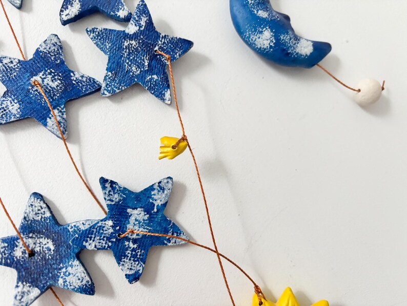 Op de afbeelding: Een blauwe en witte keramische mobiel met sterren en een halve maan die aan koperdraad hangt. De mobiel heeft gele bloemenaccenten.