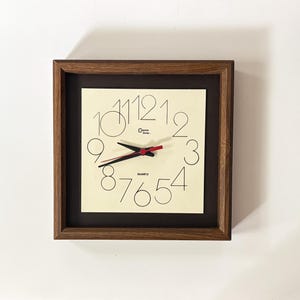 Puede incluir: Un reloj de pared cuadrado con un marco de madera marrón oscuro y una esfera de color crema. El reloj presenta números negros finos del uno al doce, manecillas negras para horas y minutos, y un segundero rojo. El texto "Jumo time" y "QUARTZ" están impresos.