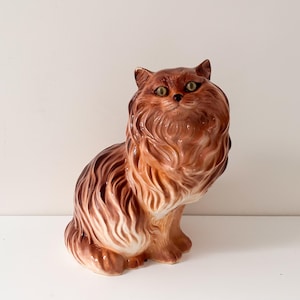 May include: A ceramic cat figurine with a fluffy, detailed brown and cream-colored coat. The cat has green eyes and is posed in a seated position. The figurine is a decorative item.