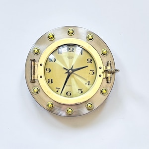 Könnte beinhalten: Eine runde Quarz-Uhr im nautischen Stil mit einem silberfarbenen Metallrahmen und einem goldfarbenen Zifferblatt. Die Uhr hat schwarze Zeiger und Ziffern sowie goldfarbene Akzente. Der Durchmesser der Uhr beträgt ca. 20 cm.