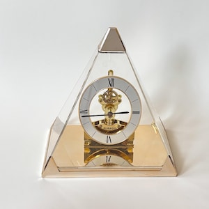 Peut inclure: Une horloge en forme de pyramide avec une base et des accents dorés, et un couvercle transparent. Le cadran de l'horloge est blanc avec des chiffres romains noirs et des aiguilles dorées. Le mécanisme de l'horloge est visible. Le mot "MIRADO" est imprimé.