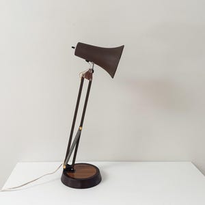 May include: A dark brown desk lamp with a flared shade, a multi-jointed adjustable arm, and a circular base featuring a wood grain inlay. The lamp includes a thin white power cord extending from the base and metallic hardware at the joints.