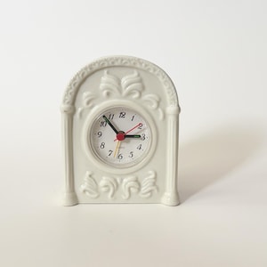 Puede incluir: Un pequeño despertador de cerámica blanquecina con una parte superior arqueada decorativa y detalles ornamentados. La esfera del reloj tiene números negros, manecillas negras y un segundero rojo. El reloj mide unos 10 cm de alto.
