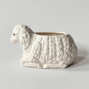 Puede incluir: Una maceta de cerámica blanca con forma de cordero. El cordero está en posición sentada con una textura de pelaje detallada y un interior ahuecado para plantar. El cordero tiene un ojo pequeño y oscuro.