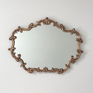 May include: A wall mirror featuring an ornate, gold-toned frame with intricate scrollwork and carved detailing. The mirror has a distinctively shaped, irregular outline with symmetrical curves and flourishes along the entire perimeter.