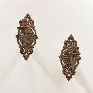 May include: A pair of wall-mounted candle holders featuring an ornate, symmetrical scrollwork design in a dark bronze finish. Each piece includes a circular platform for a candle, integrated into a decorative, elongated, diamond-shaped metal frame.