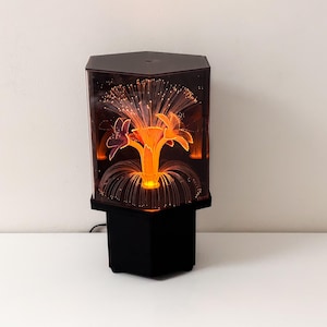 Pode incluir: Uma lâmpada decorativa com base preta e topo hexagonal. A lâmpada apresenta um design floral laranja central com fios de fibra ótica que emitem um brilho quente. A lâmpada está sobre um fundo branco.
