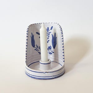 May include: A white ceramic wall-mounted candle holder with a semi-circular back panel featuring hand-painted blue floral motifs and striped edges. A single white taper candle is positioned in the central circular base of the holder.