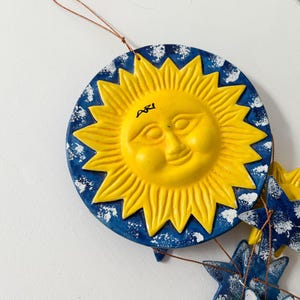 Op de afbeelding: Een blauwe en gele keramische zon met een glimlachend gezicht en het woord "Agi" erop geschreven. De zon maakt deel uit van een mobiel met blauwe en witte sterren.