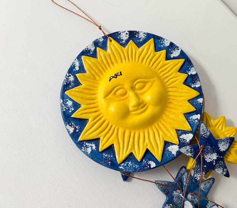 Op de afbeelding: Een blauwe en gele keramische zon met een glimlachend gezicht en het woord "Agi" erop geschreven. De zon maakt deel uit van een mobiel met blauwe en witte sterren.