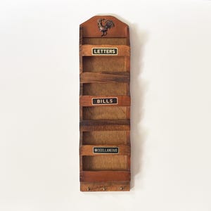 May include: A vertical wooden wall organiser featuring three tiered slots labelled with black and white plaques reading LETTERS, BILLS, and MISCELLANEOUS. The top section displays a carved rooster motif, and the base includes three small metal hooks.