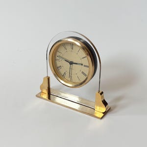Könnte beinhalten: Eine goldfarbene Tischuhr mit cremefarbenem Zifferblatt und schwarzen römischen Ziffern. Die Uhr hat einen goldenen Rahmen und eine klare, gewölbte Oberseite. Der Sockel ist rechteckig und ebenfalls goldfarben. Die Uhr ist ein dekorativer Artikel.