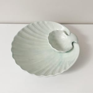 Puede incluir: Un plato de servir de cerámica blanca con forma de concha marina con un tazón más pequeño adjunto. El plato tiene un esmalte azul claro.