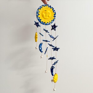 Op de afbeelding: Een gele zon met een gezicht en blauwe sterren die aan een touwtje hangen. De zon en sterren zijn gemaakt van keramiek.