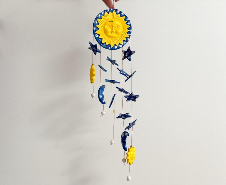 Op de afbeelding: Een gele zon met een gezicht en blauwe sterren die aan een touwtje hangen. De zon en sterren zijn gemaakt van keramiek.