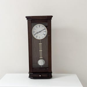 May include: A tall, rectangular wall clock with a dark brown wooden frame and fluted side columns. The clock face is white with black Roman numerals and hands. A silver-toned pendulum hangs behind a glass panel, above a small bottom drawer.