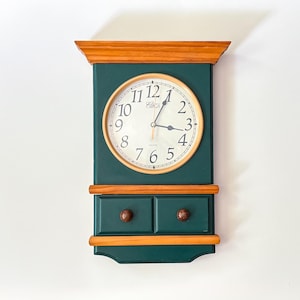 May include: A wall clock with a dark green rectangular wooden frame and natural wood accents. The clock face is round with black Arabic numerals and the text "Cuckoo" printed in the centre. Below the clock are two small drawers with round knobs.